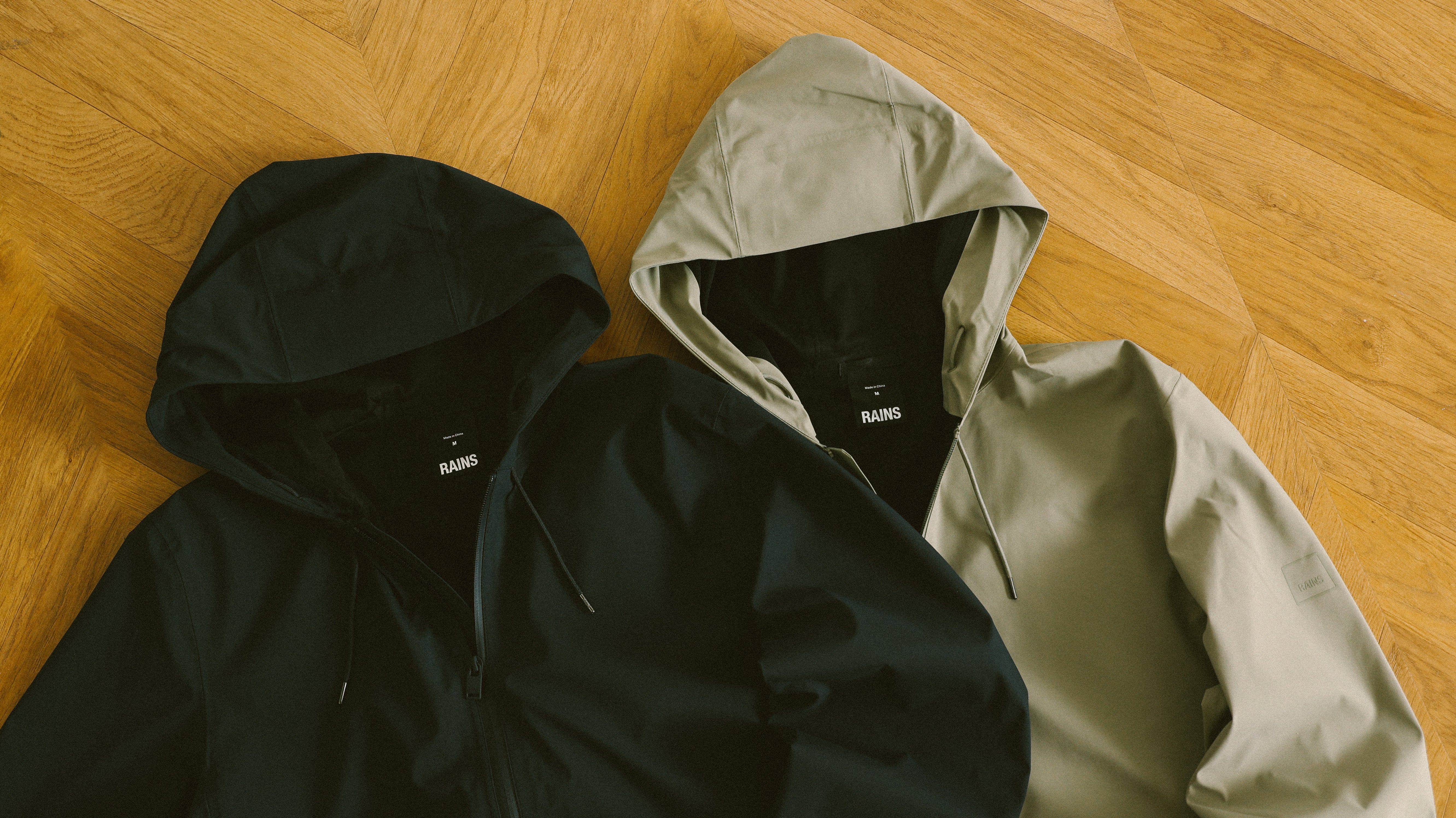 Two Rains rain jackets, one black and one beige, both with hoods, laid out on a wooden herringbone floor.