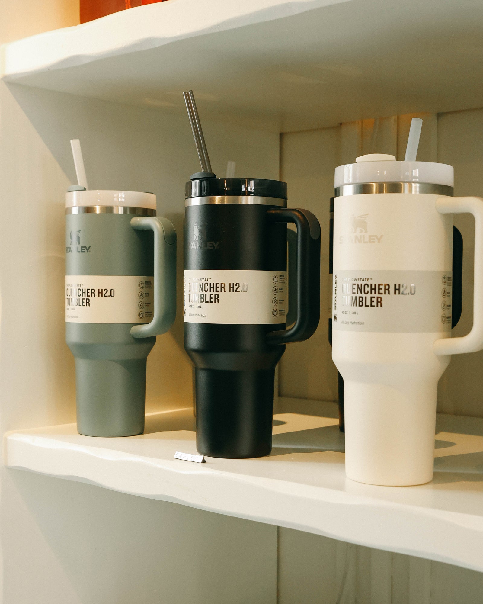 Three Stanley tumbler cups with handles and straws on a white shelf. The logo is centered on each cup. Colors from left to right: sage green, black, and white.