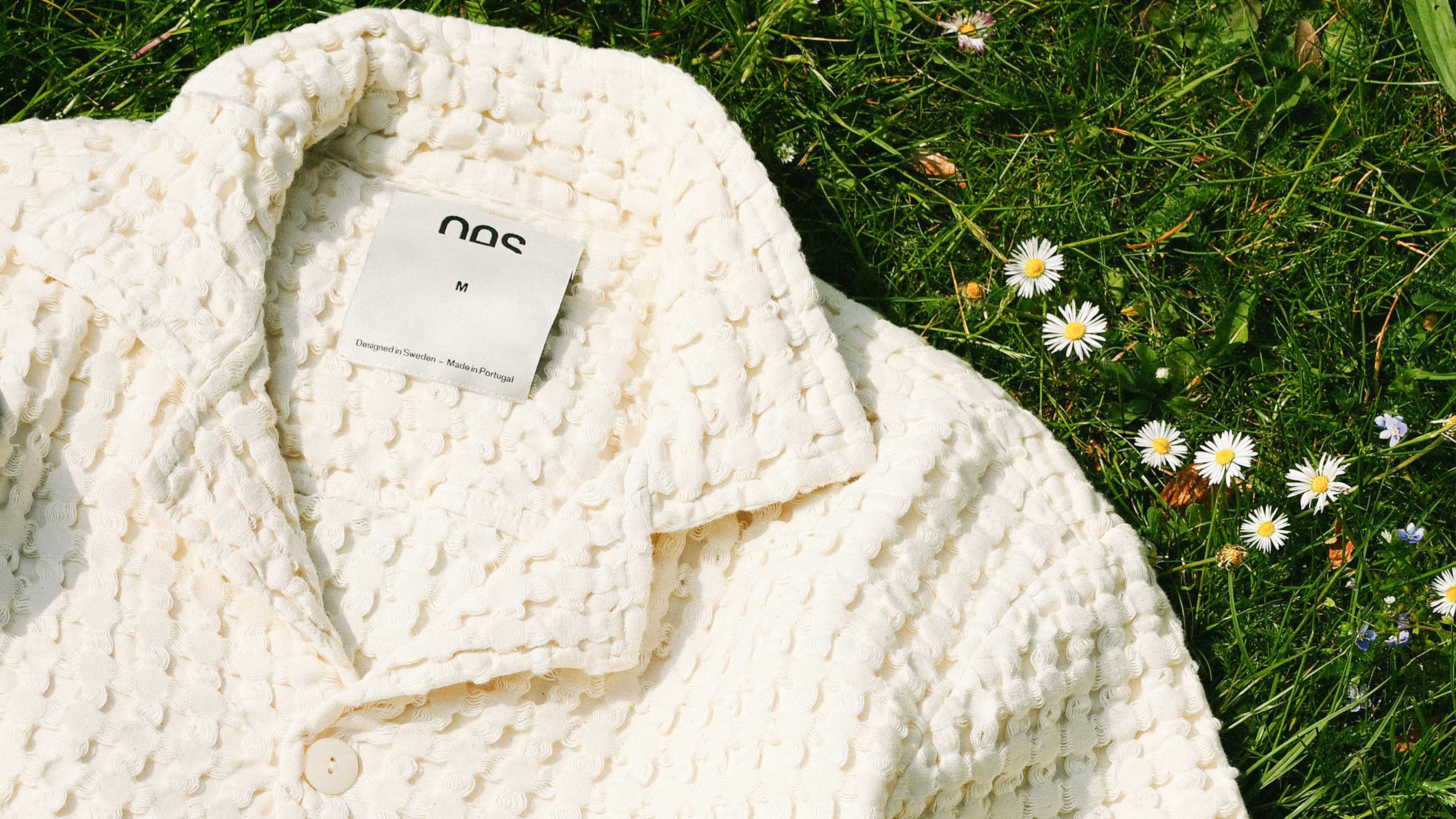 White textured OAS blouse lying in grass with daisies. The collar, shoulders, and top button area of the shirt are visible.