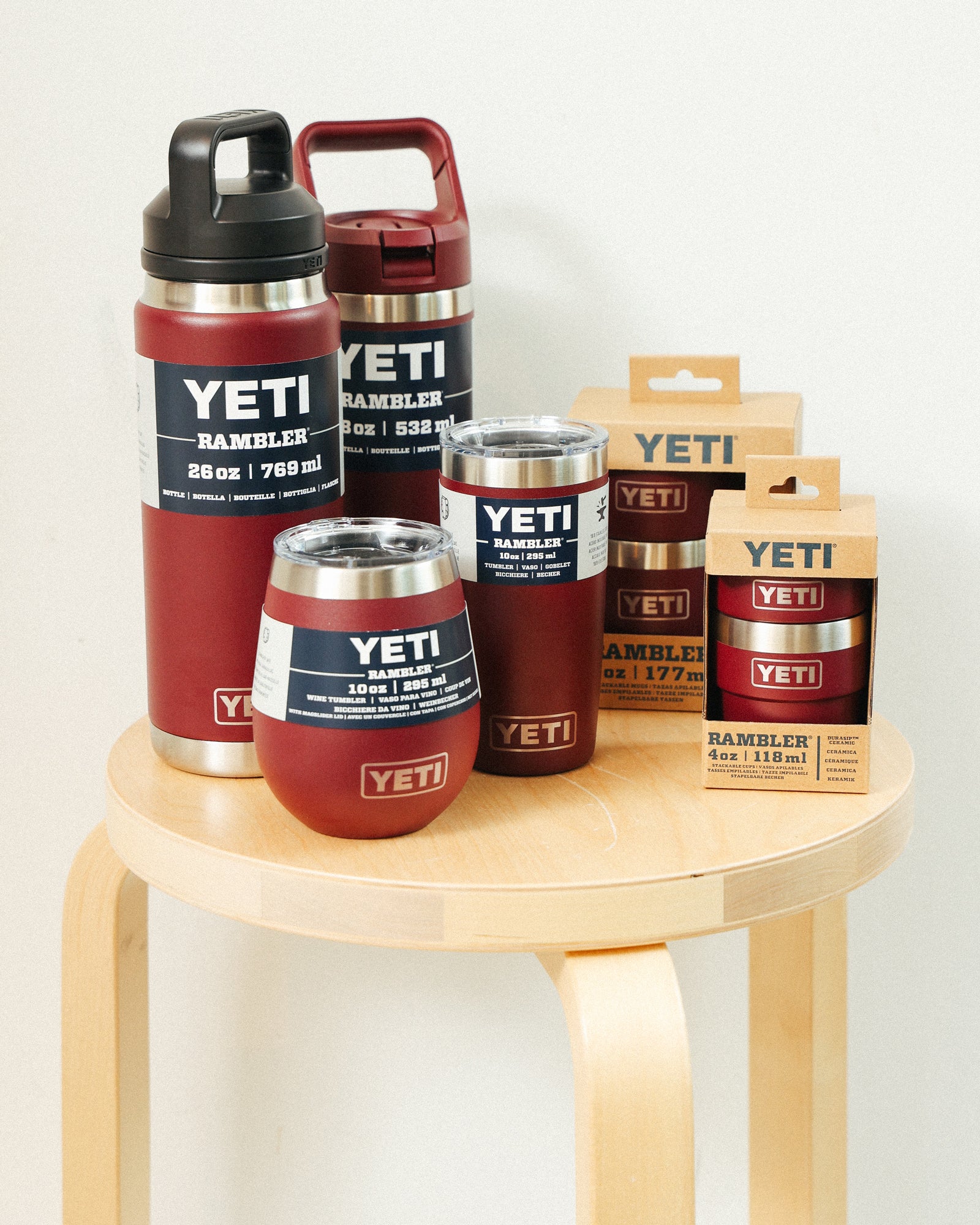 Full Yeti tumbler collection in burgundy red on a wooden stool. Many tumblers mix metal and red colors, one has black and red. All are still in their packaging. Light background.