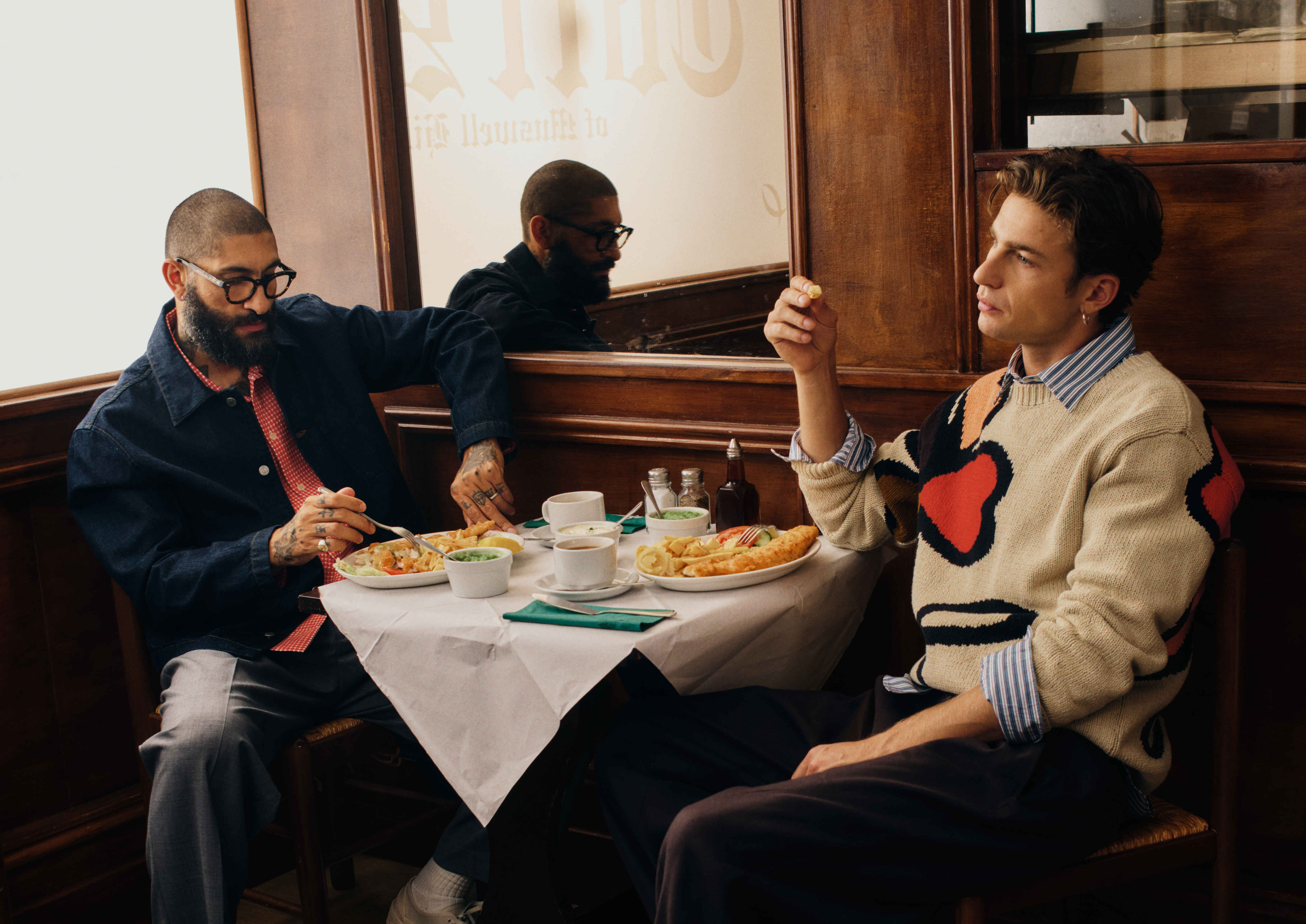 A brown NN07 sweater with the logo in a rope-like design on the chest, photographed in front of a red garage door.