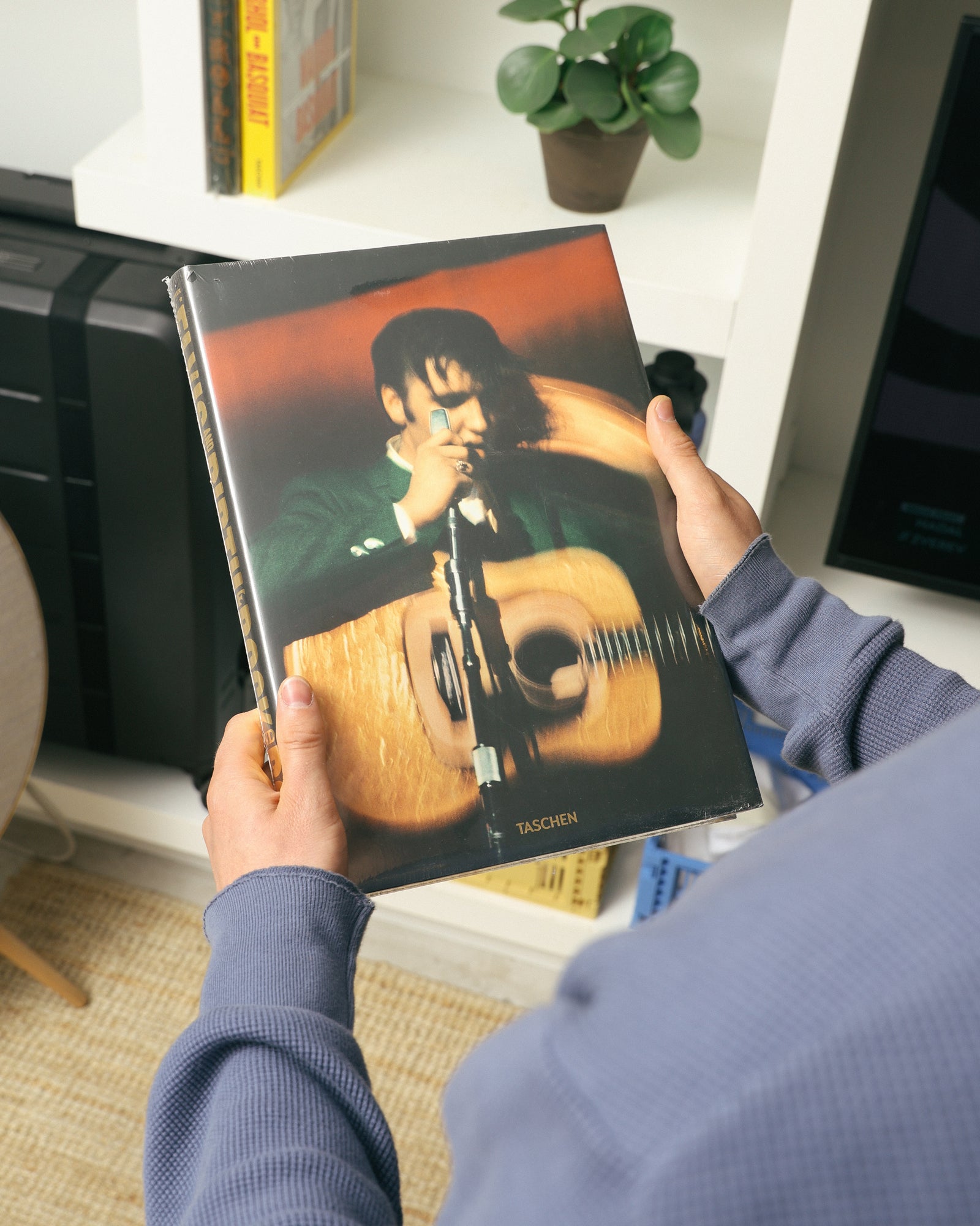 Person holding a book, seen over the shoulder. Wearing a faded blue sweater. The book cover shows a person with a guitar and the word 'Taschen'. In the background: a white cabinet with a plant, crates, and two books.