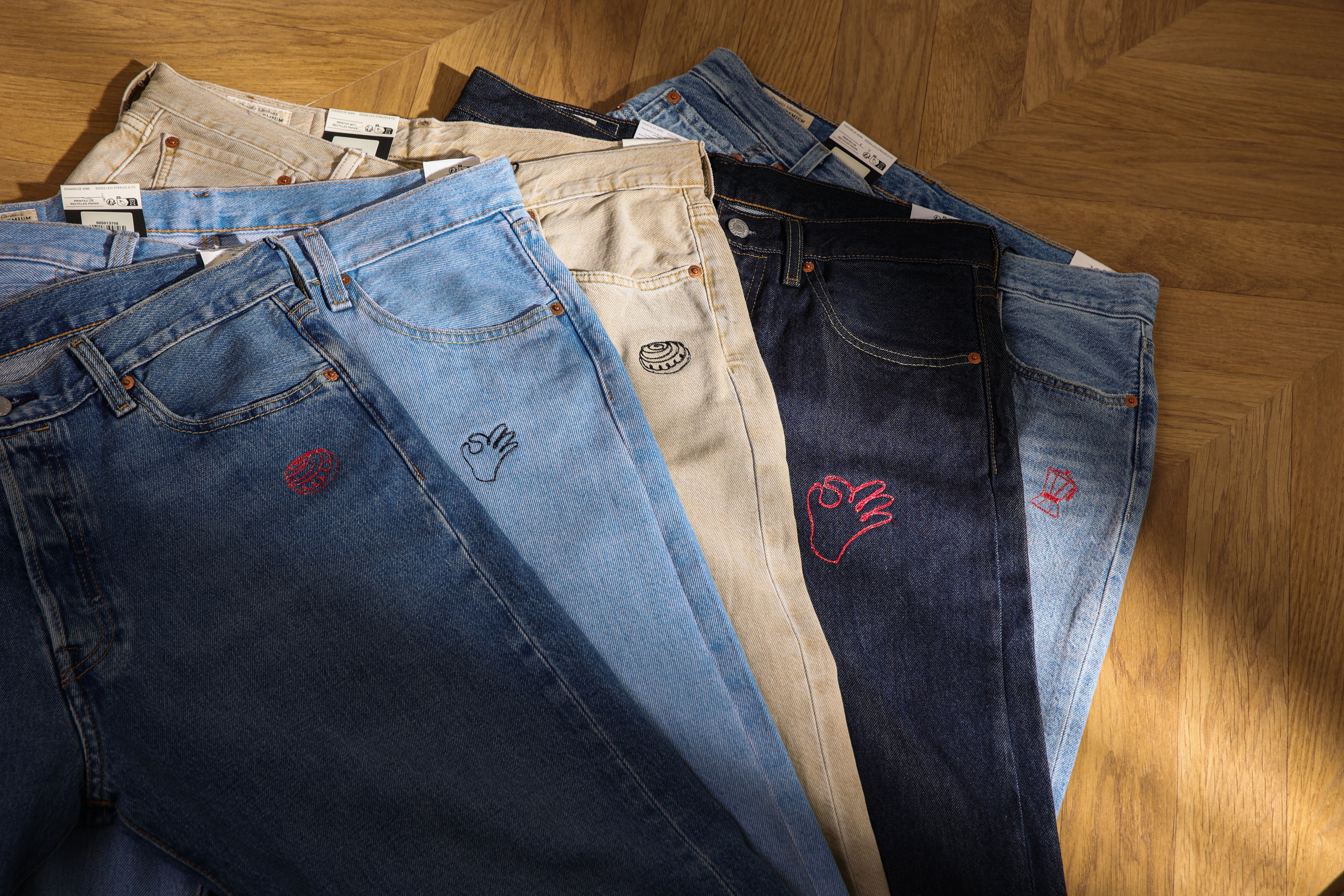 Five Levi’s jeans in different shades of blue and cream laid out on a wooden herringbone floor. Each has small embroidery including a cinnamon bun, an OK hand sign, another cinnamon bun, another OK hand, and a coffee pot.