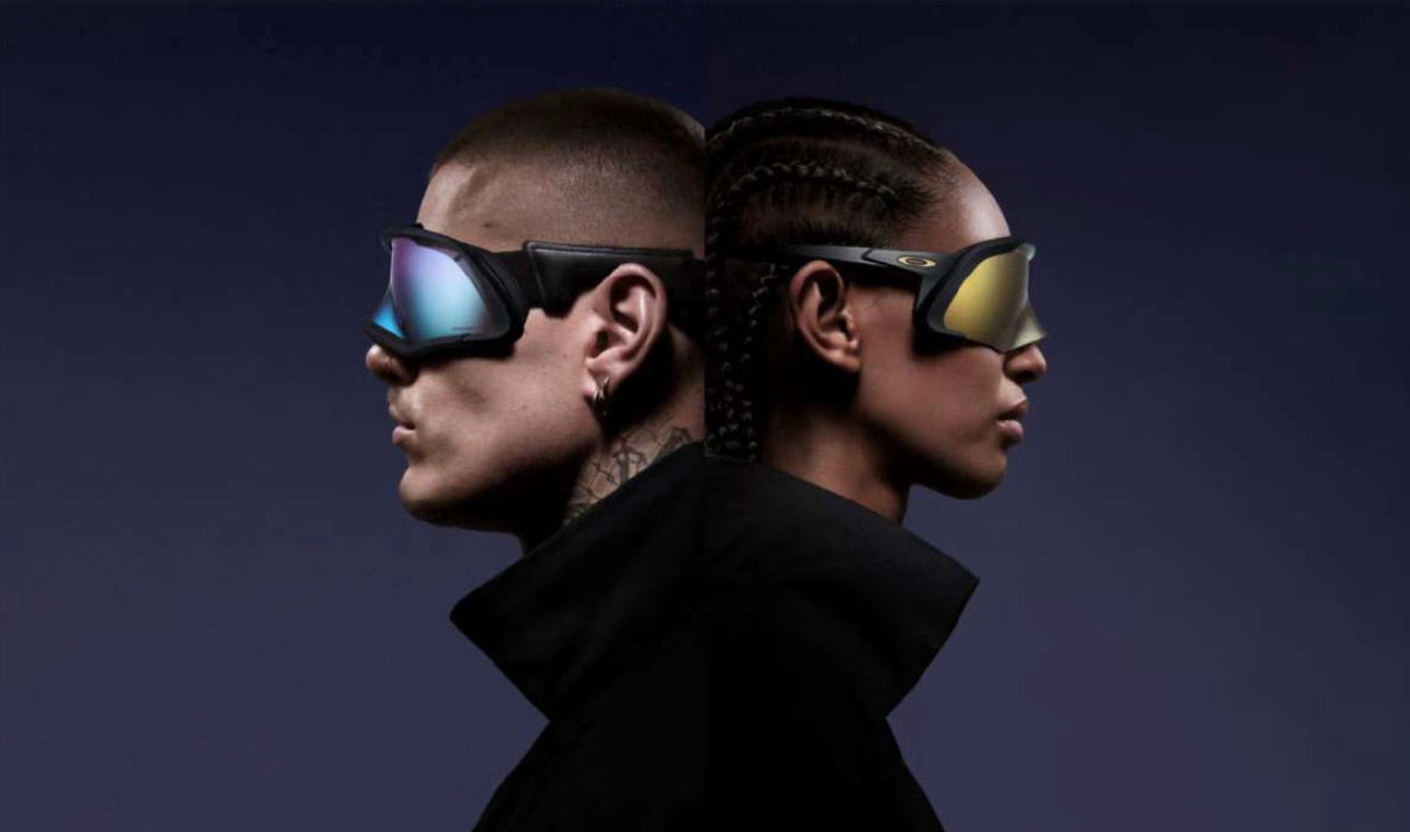 Two people dressed in black, seen from head to shoulders, standing back to back. Both wear large Oakley sunglasses, one gold matte with black frame and the other with a purple gradient and black frame, against a dark background.
