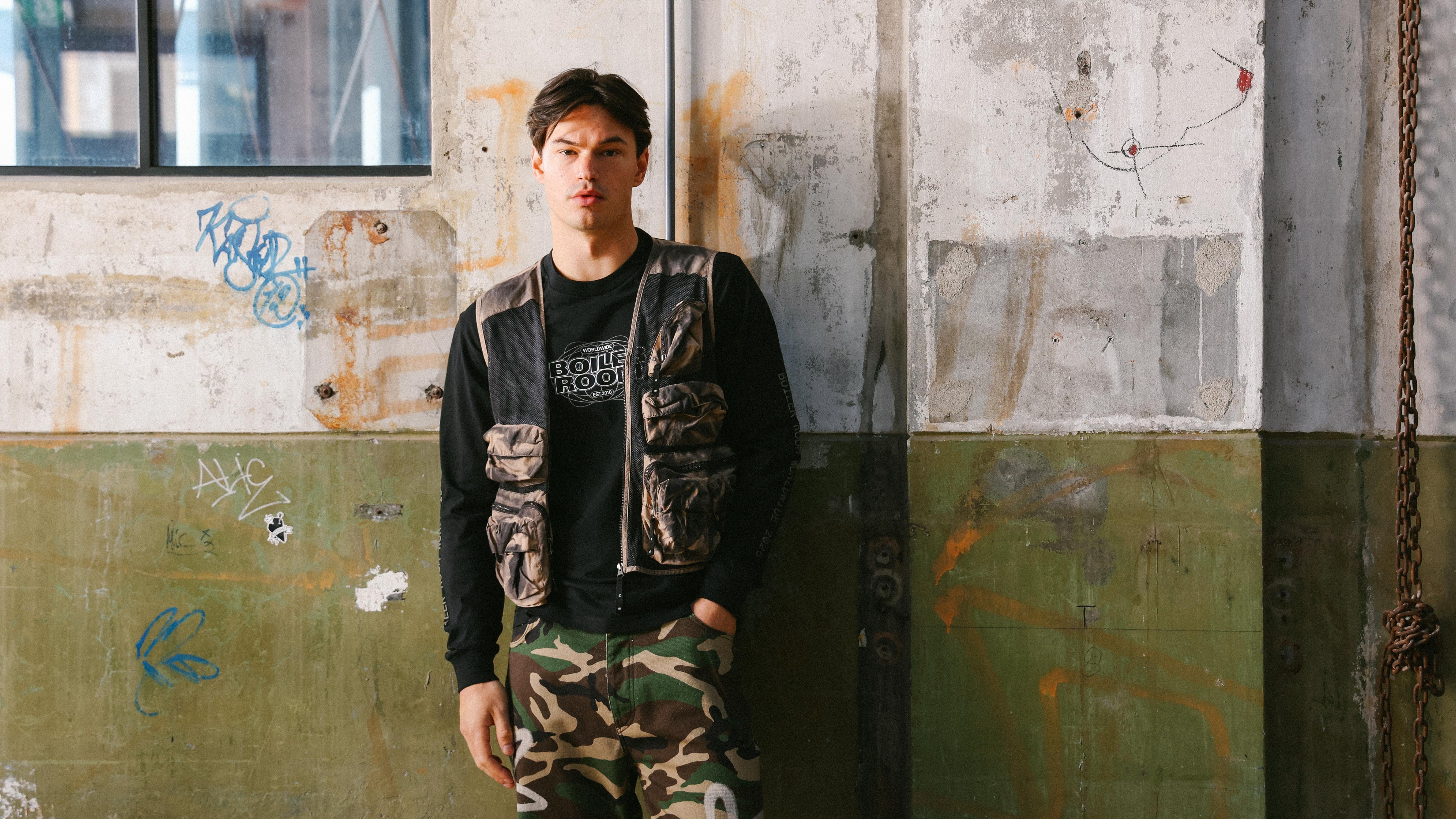 A man stands in front of a white-green concrete wall with graffiti. A rusted chain hangs to the right. He wears a Boiler Room sweater under a brown vest with black details and pockets, camo cargo pants, and has one hand in his pocket