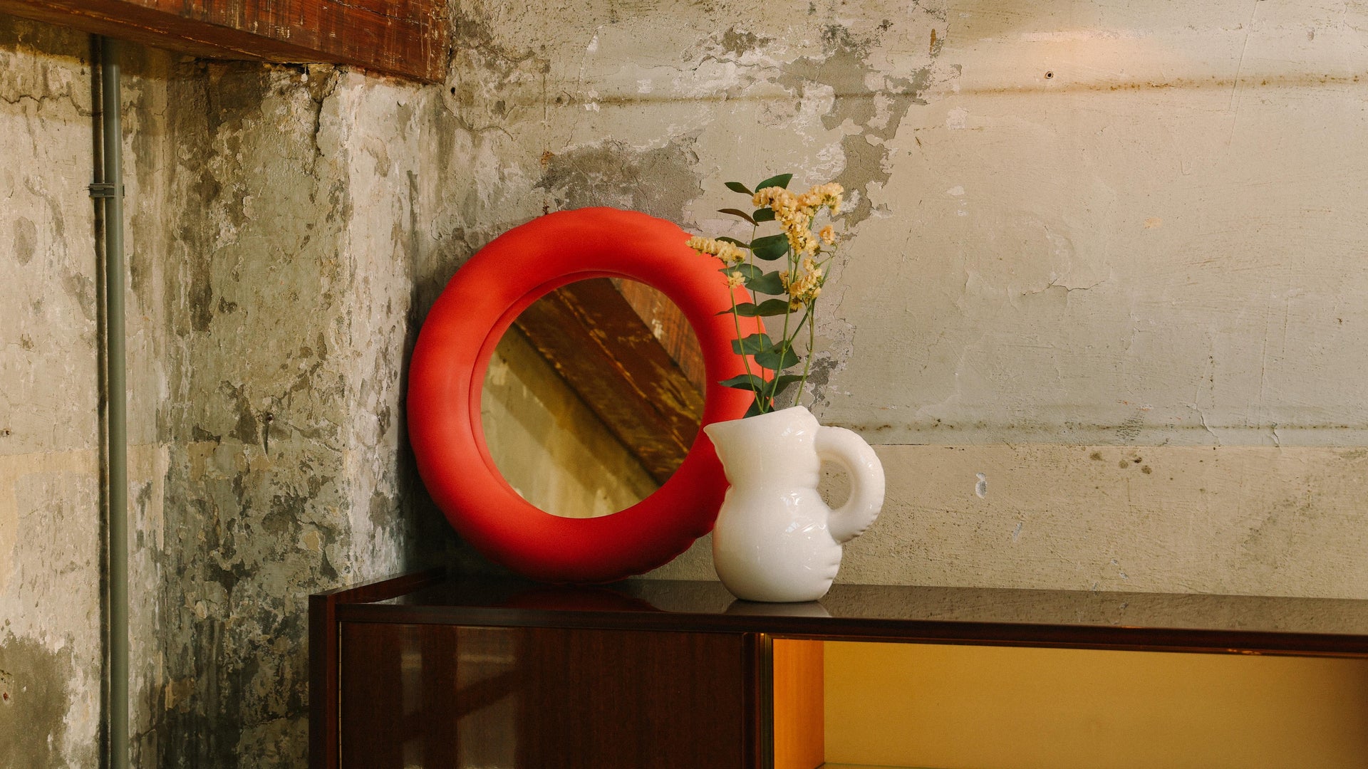 A wooden cabinet against a concrete wall, topped with a round red mirror with a thick frame and a white pitcher-shaped vase holding flowers.