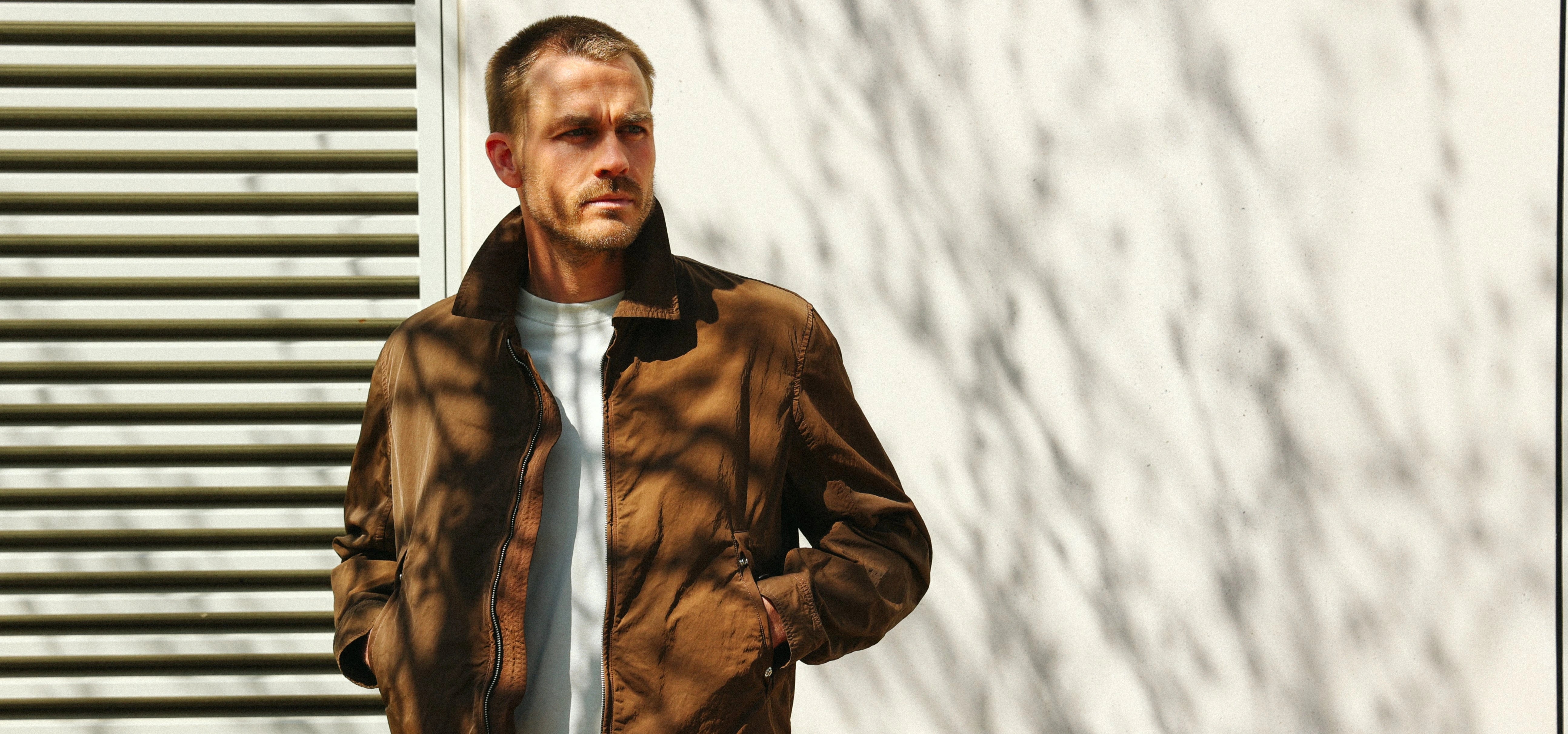 Man wearing a brown jacket with the collar up and a white T-shirt underneath. The background is a white wall with a window, and shadows of tree branches are visible.