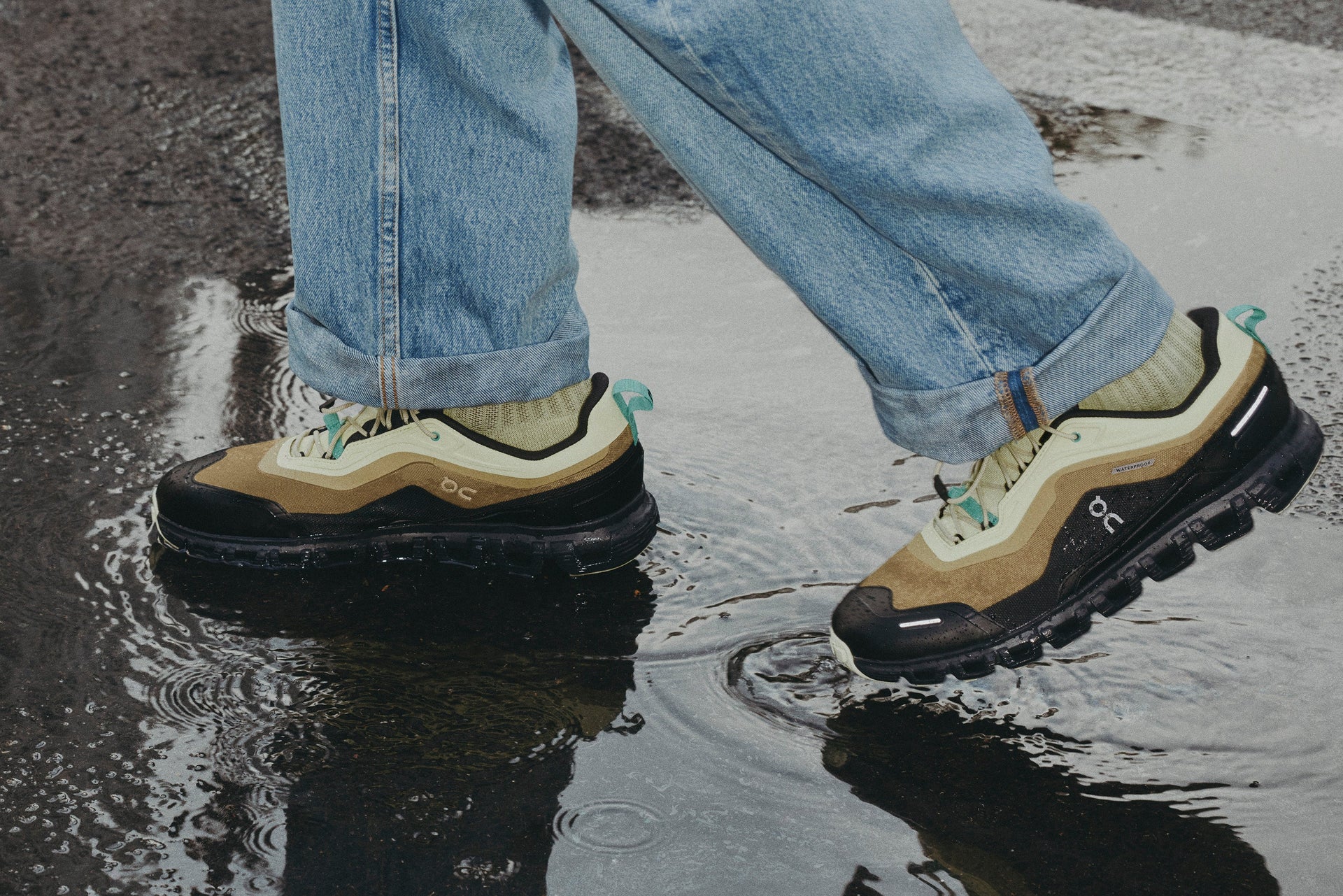 Four ON running shoes on a stone floor with a person standing between them wearing one intact shoe and one shoe cut in half, showing white socks.