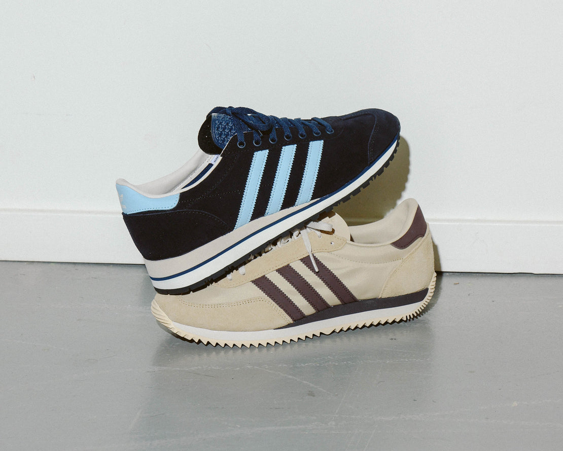 Two pairs of Adidas sneakers stacked on a gray floor with a white wall background.